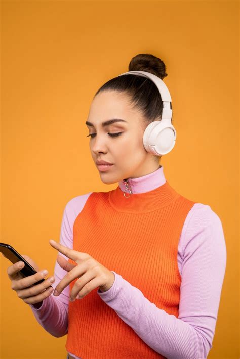a woman with headphones on looking at her cell phone while wearing an orange sweater