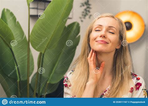 Blonde Woman in Living Room Next To Tall Plant Looking Relaxed and Smiling Holding Hand To Her ...