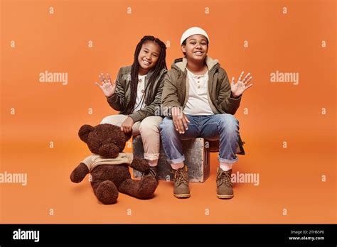 jolly preteen african american children in winter outfit posing with teddy bear and waving at ...