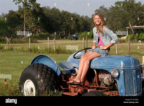 A beautiful blond girl sitting on a tractor in the middle of a farm ...