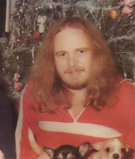 a man with long hair holding two small dogs in front of a christmas tree and looking at the camera