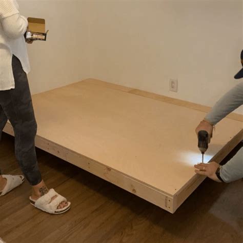 two people working on a bed frame in a room with hard wood floors and white walls