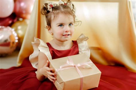 Closeup of Two Year Old Baby Girl in a Red Dress Holding Her Birthday Gift and Looking at Her ...