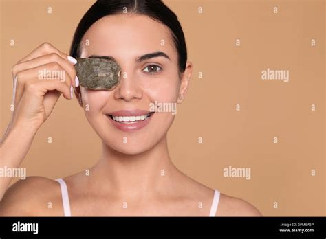 Beautiful young woman putting green tea bag on eye against beige background, space for text ...