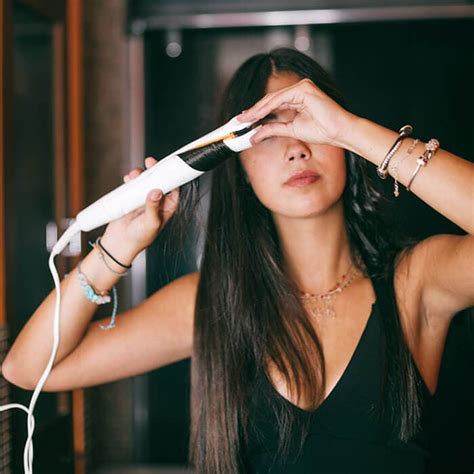 Brunette young woman styling her long hair with curling iron or ...