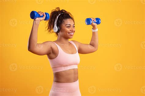 A young woman in sportswear lifts dumbbells while smiling. She wears