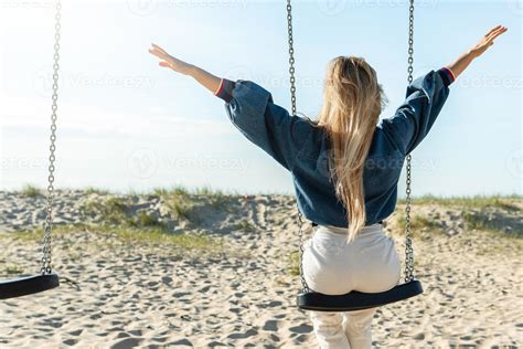 Rear shot of young woman swinging on swing and spreading hands at the