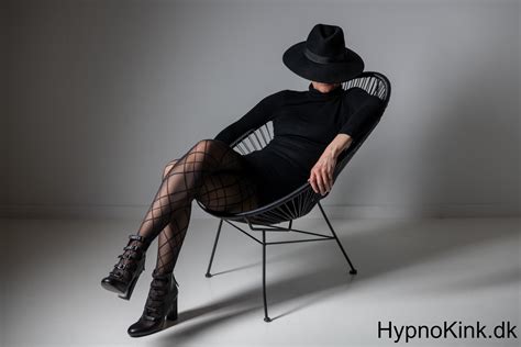 Hypno Mistress i sort kjole og sorte strømper, i en sort hat sidder i