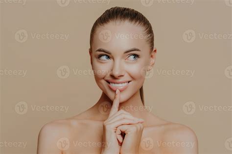 Portrait of good looking woman looks aside mysteriously, keeps finger