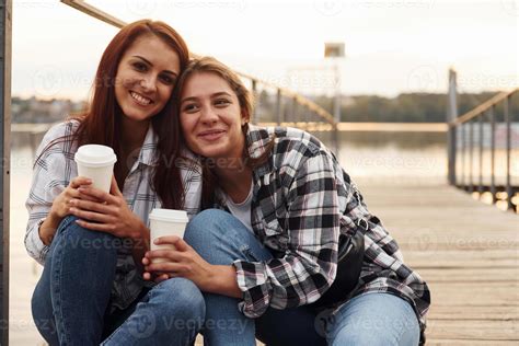 Cute lesbian couple sits together near the lake with cups of drink in